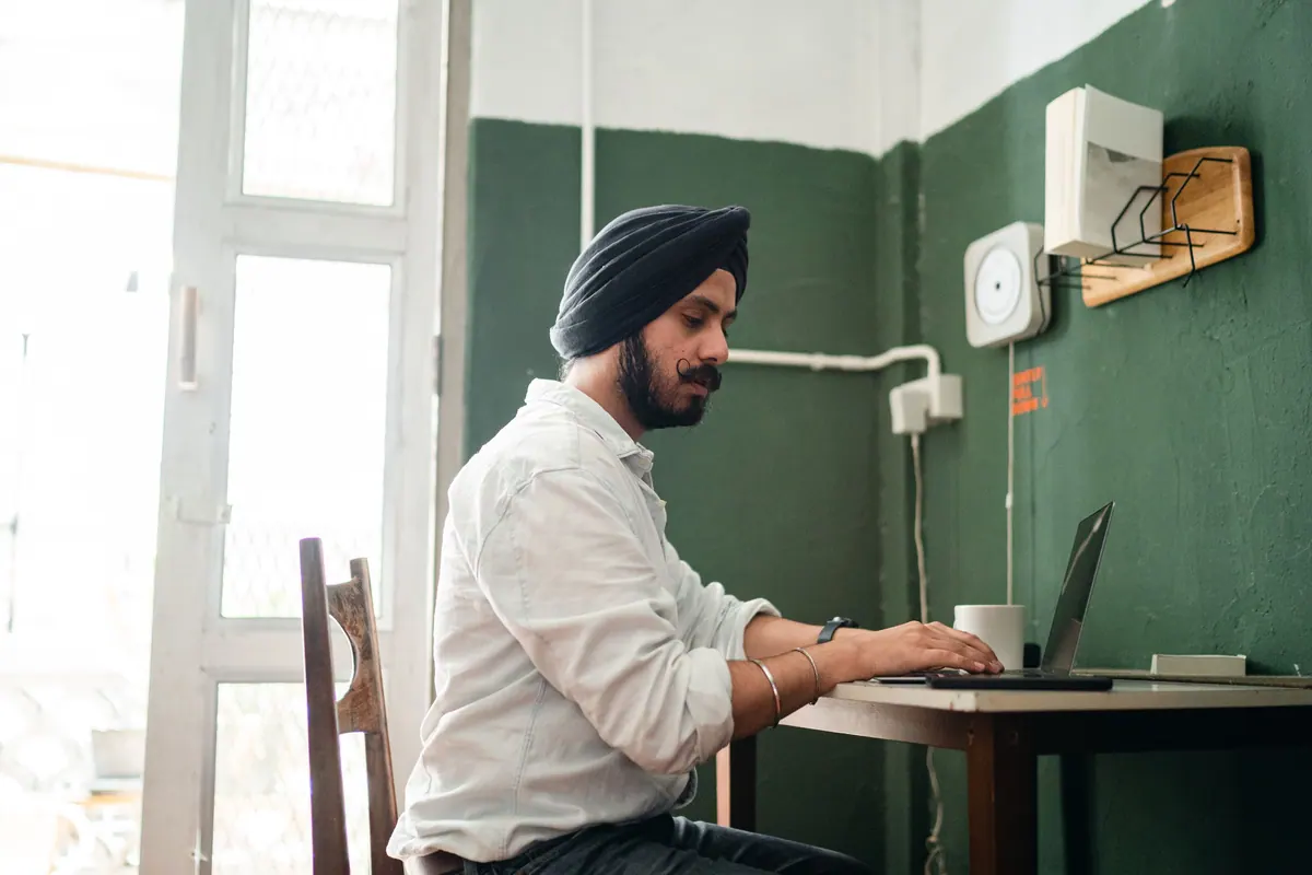 A man in a turban working at a laptop on a small table in a plain green-walled office