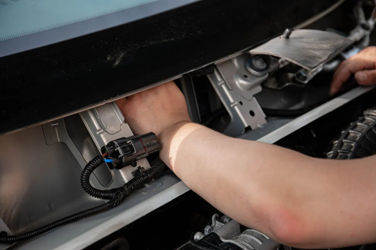 A hand reaching behind a vehicle panel to a wiring-harness connector