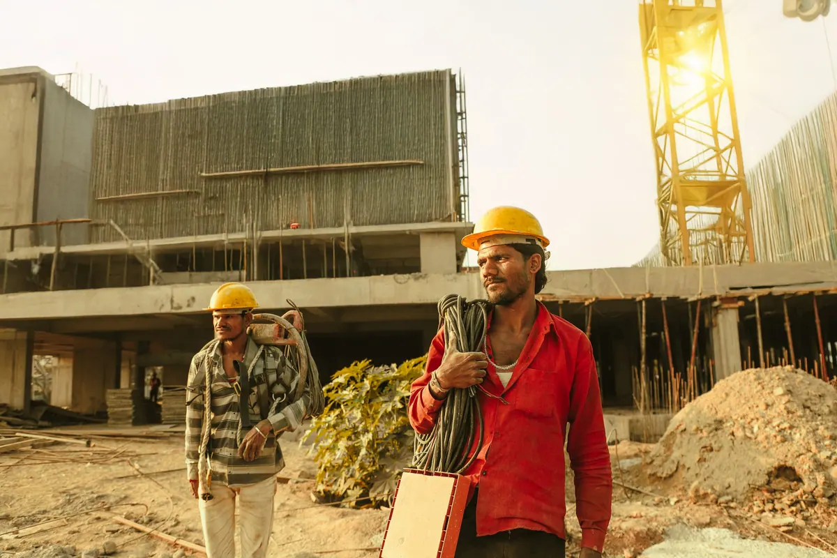 Two construction workers in helmets at a building site with a tower crane behind
